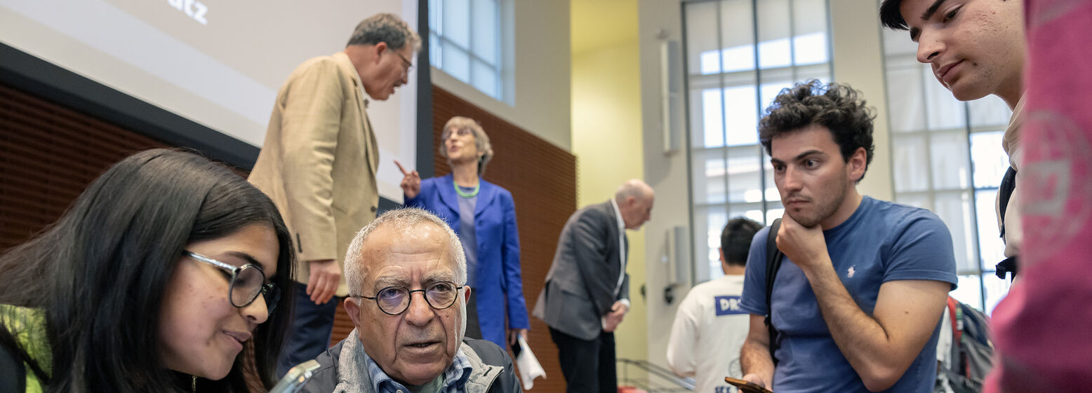 An older man sits at the edge of a stage and talks with several students. Behind him, a tall man talks with a shorter woman, and a middle-aged man bends forward to speak to students in front of him.
