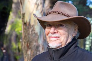 Paul David in his older years wearing a wide-brimmed brown hat and standing among some trees