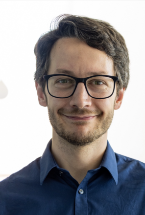 Matteo Ippoliti smiling without teeth and wearing a medium-blue collared shirt and wearing black-rimmed glasses