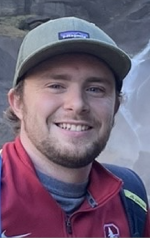 Jesse Hoke wearing a sage-green baseball cap, a red Stanford zip-up sweater, and a hiking backpack and standing in front of a rocks and mist from a waterfall