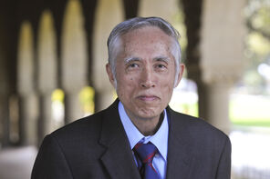 Tze Leung Lai wearing a light-blue collared shirt, royal blue and red tie, and dark coat standing under one of the arcades along Stanford University's Main Quad