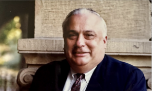 Walter Falcon wearing a navy-blue coat, white collared shirt, and a dark tie in front of a sandstone pillar 