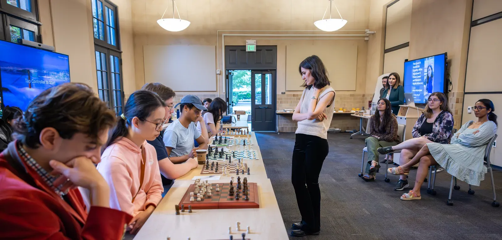 A woman stands in front of a long table with six chess boards and six seated players