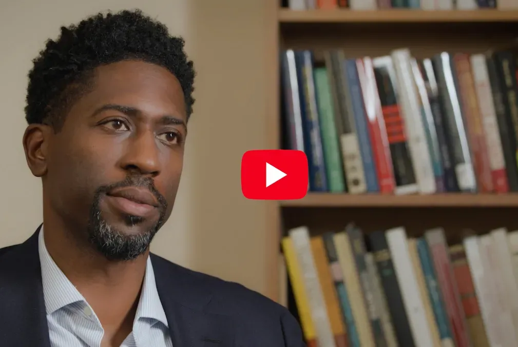 Image of Lerone A. Martin, a Black man wearing a collared shirt and a sport coat in his academic office next to a full bookshelf